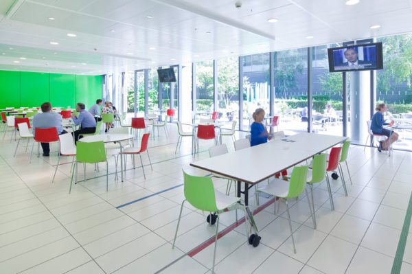 lunching area with television on, informal chats going on round tables and alternating red and green chairs