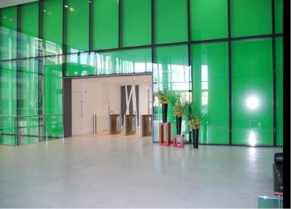 bright green glass panelled wall with opening allowing access through entrance barriers, tall vases of flowers to the side
