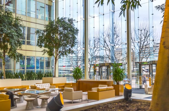 ground floor business lounge with mustard and brown furnishing, indoor plants and trees, high ceilings and view of buildings through the glass