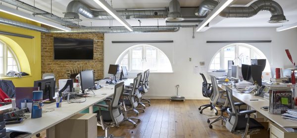 long desks of work stations in use, wooden floors and crescent shaped downs with a screen mounted on a side brick wall