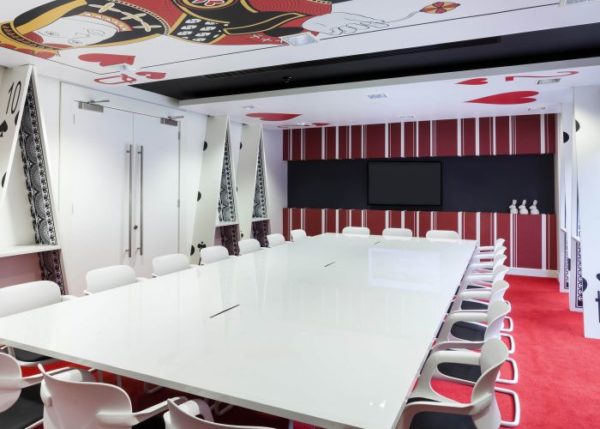 Alice in Wonderland style meeting room with white table and chairs, red carpet, white 'house of cards' walls, white rabit figurines and Queen of Hearts and Two of Hearts cards on the ceiling